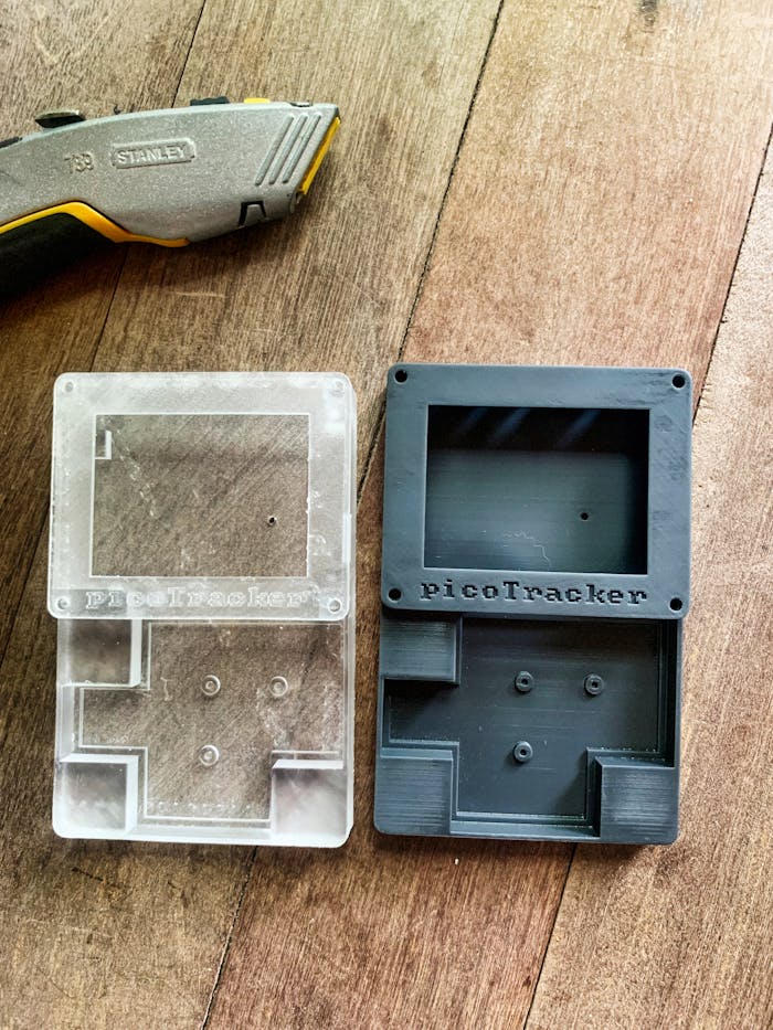services-01 Display of transparent and black 3D printed PicoTracker cases next to a utility knife on a wooden surface.